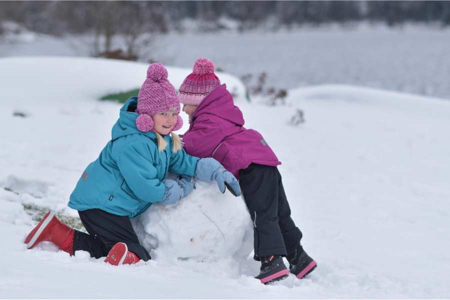 Dvě malé holčičky si hrají ve sněhu, opírají se o velkou sněhovou kouli